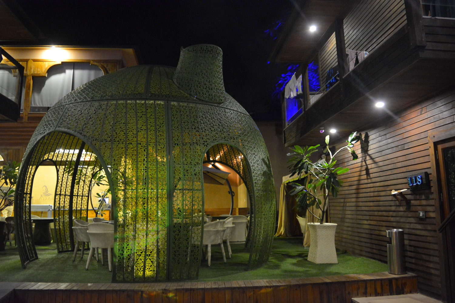An open air restaurant with tables and chairs with one table covered with a teapot shaped netted walls and dome lit at night with lamps