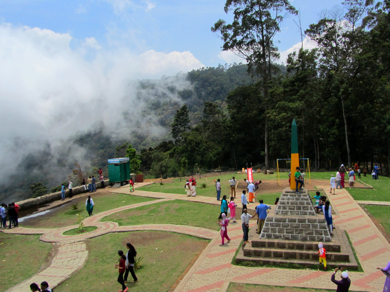 A small park with pavements, a monument, and many visitors with a hill behind it partially covered in mist