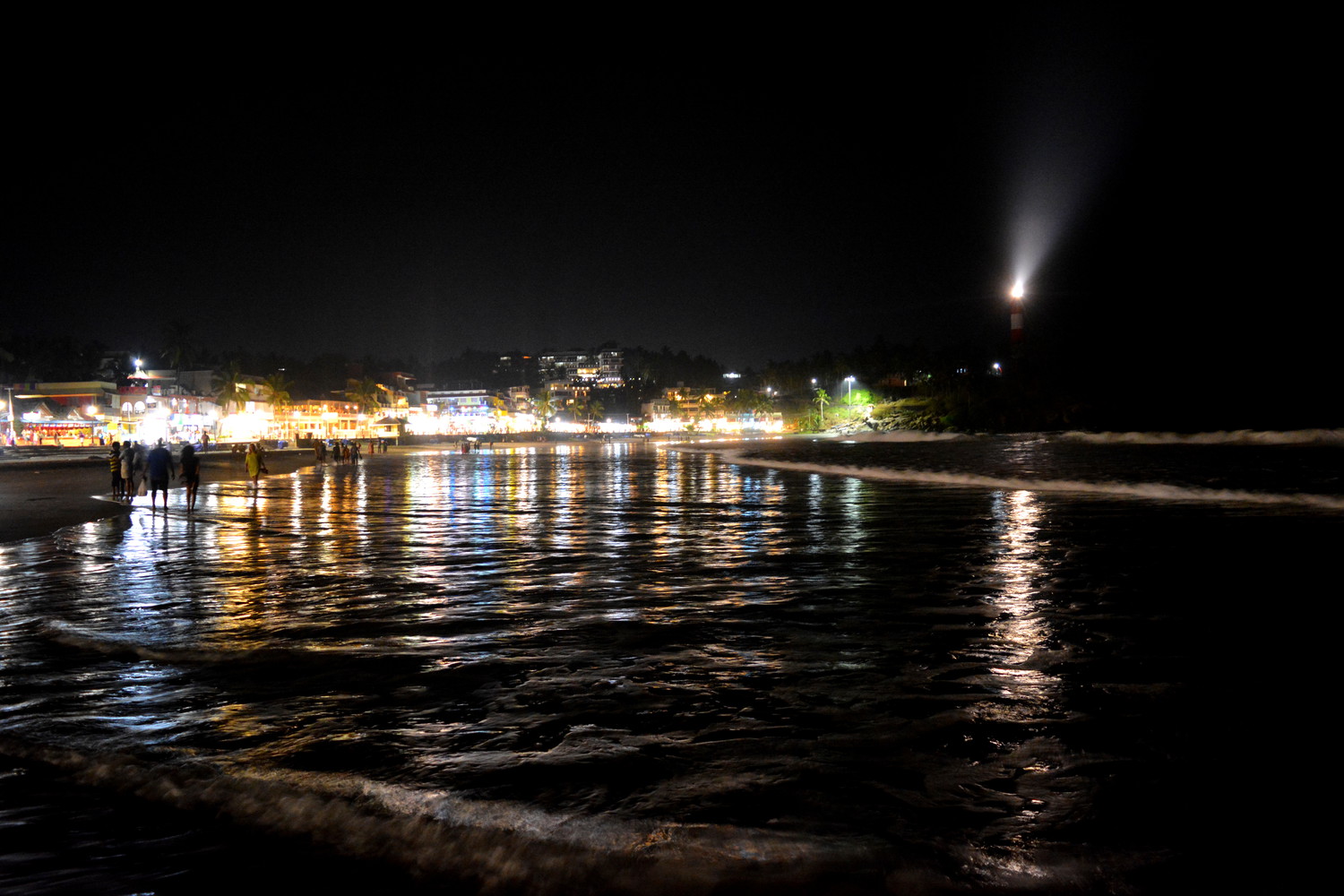 Sea beach at night with the lights from many shops and lighthouse emitting light visible in the background