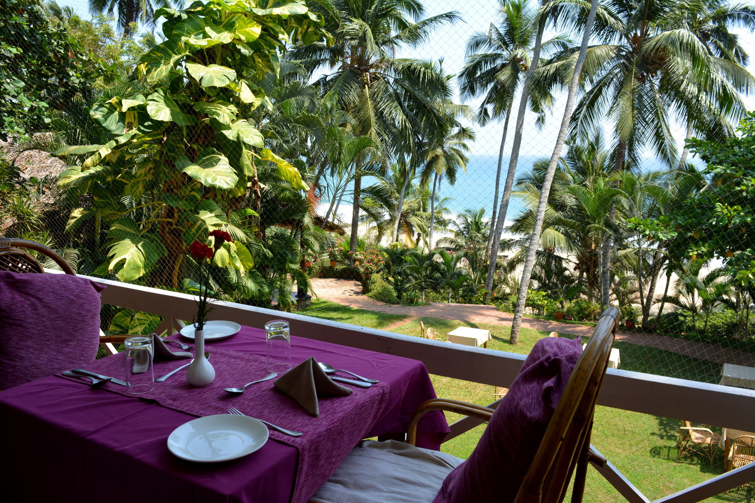 A dining table at the edge of a balcony overlooking green lawns, coconut palm trees, and sea beach below