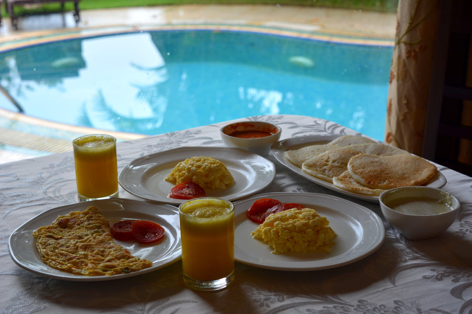 Breakfast laid out on a table with white table cloth beside a swimming pool; there are one plate of omelette served with tomato slices, two plates of scrambled eggs served with tomato slices, a plate of dosas (Southern Indian pancakes), two bowls of chutney (Indian sauce), and two glasses of pineapple juice
