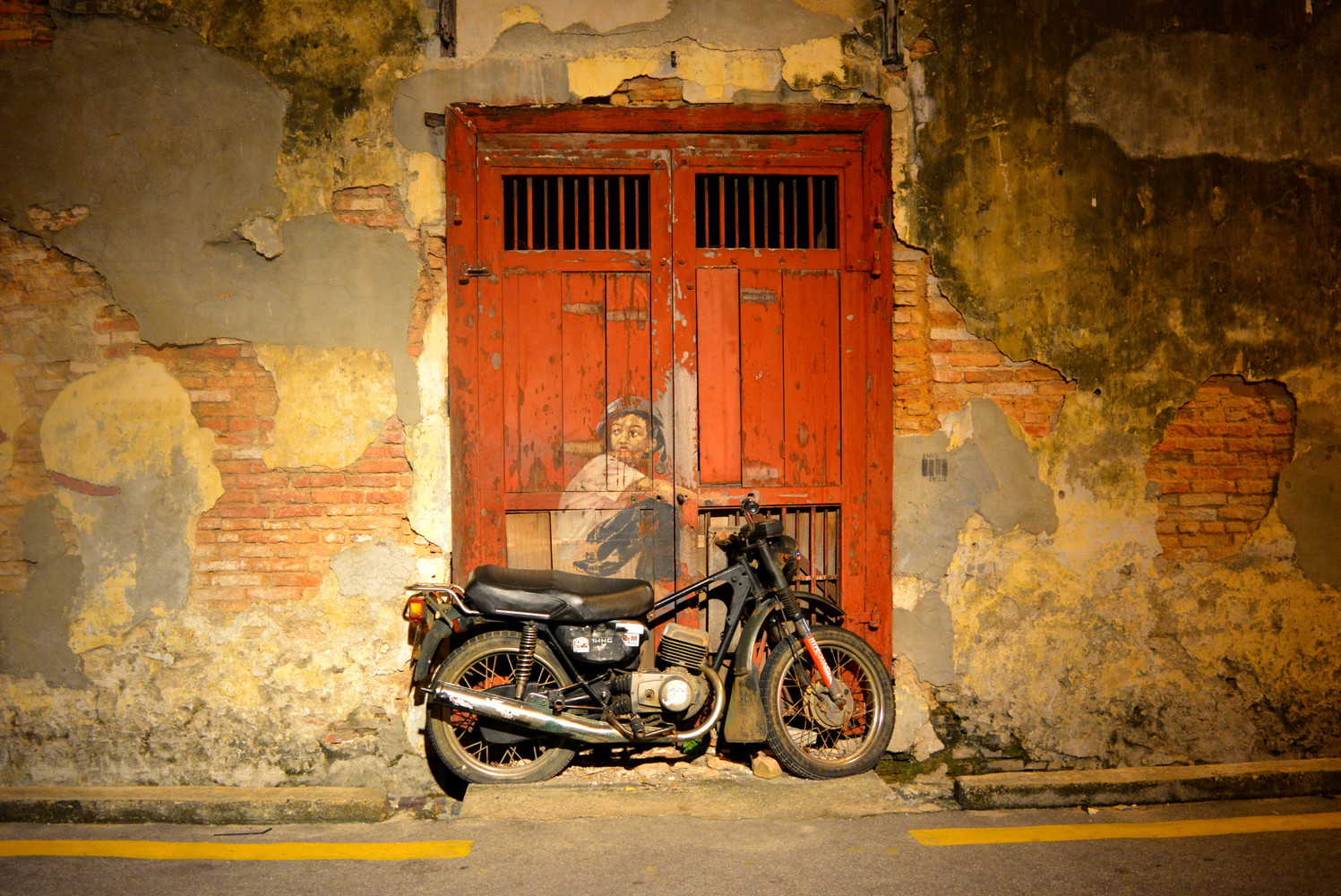 A mural with a boy painted on a door and an actual physical motorcycle attached to the door such that the painted boy appears to be sitting on the motorcycle; the wall around the door is tattered with its paint peeling off