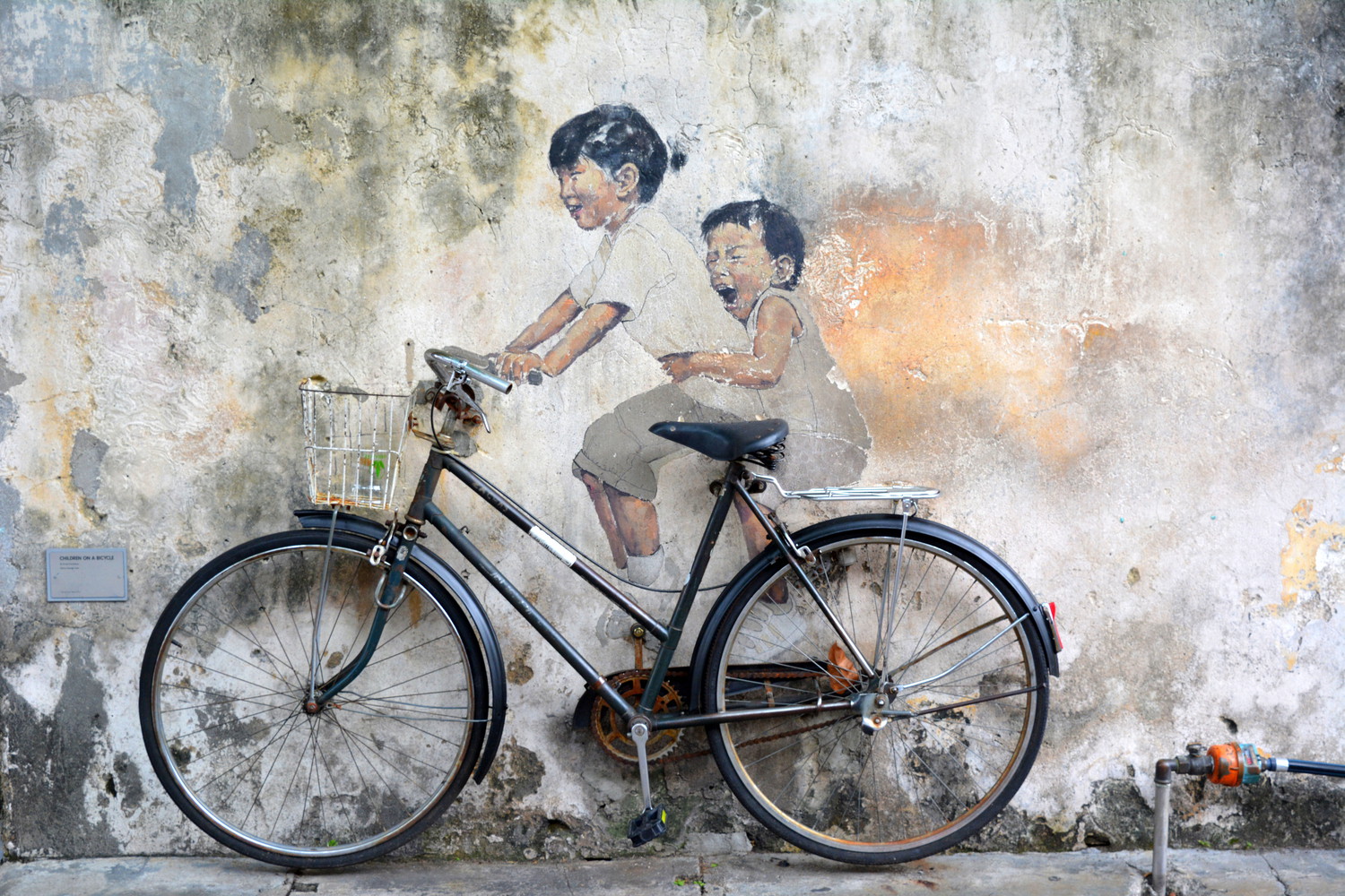 A mural with a little girl and an even smaller boy painted on a wall with an actual physical bicycle attached to the wall such that the painted children appear to be sitting on the bicycle with the girl riding the bicycle and the boy sitting on the back seat