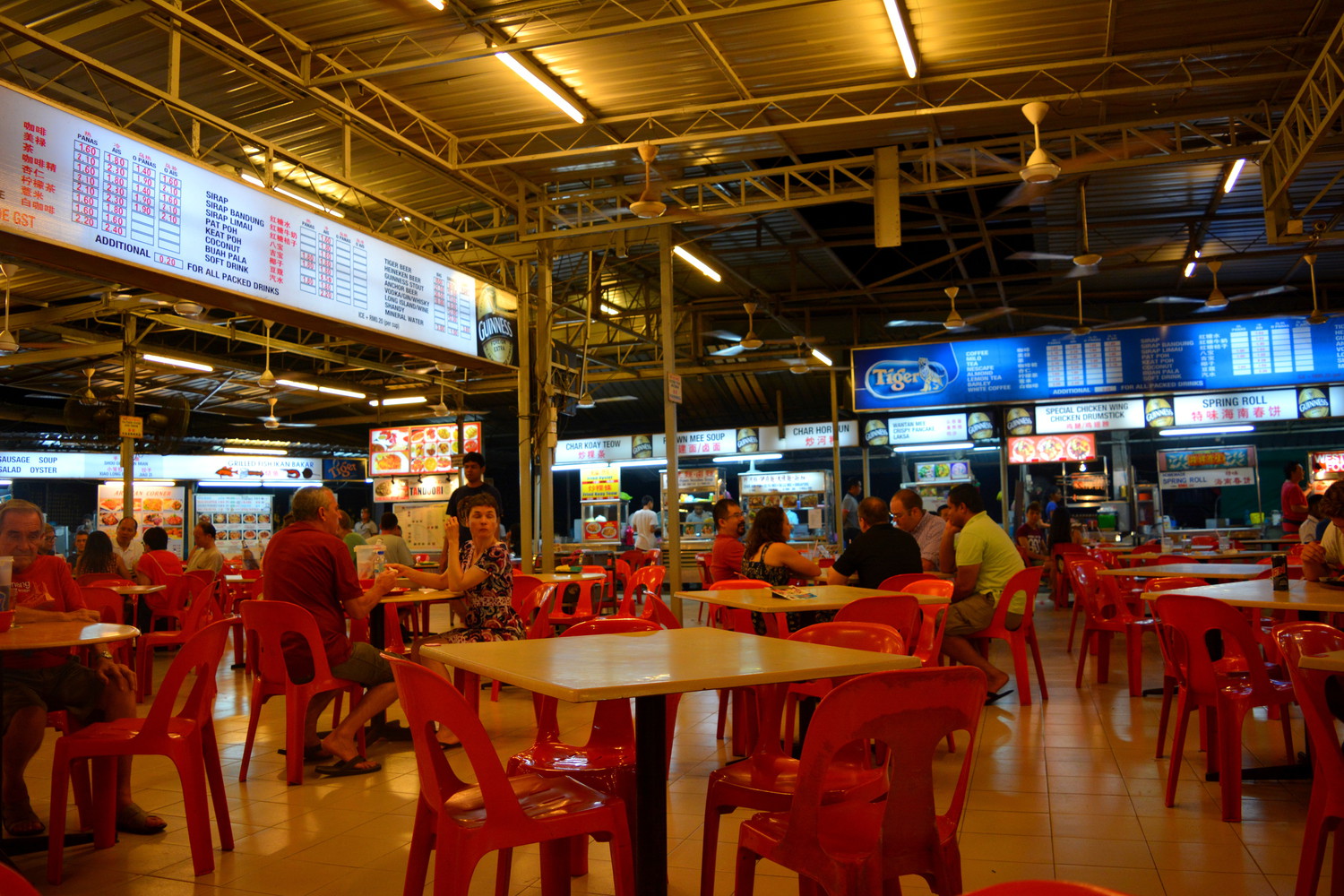 The interior of a food court with chairs, tables, and guests, and large menu posters near the ceiling