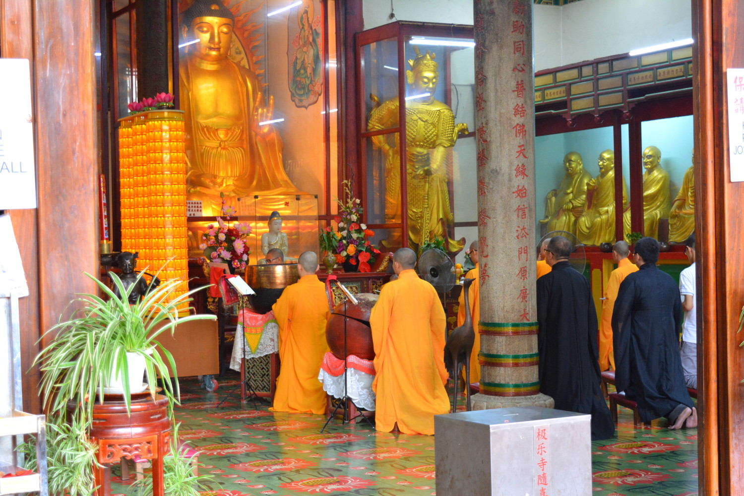 Monks in saffron and black attire sitting on their knees and praying in front of a large statue in a prayer hall