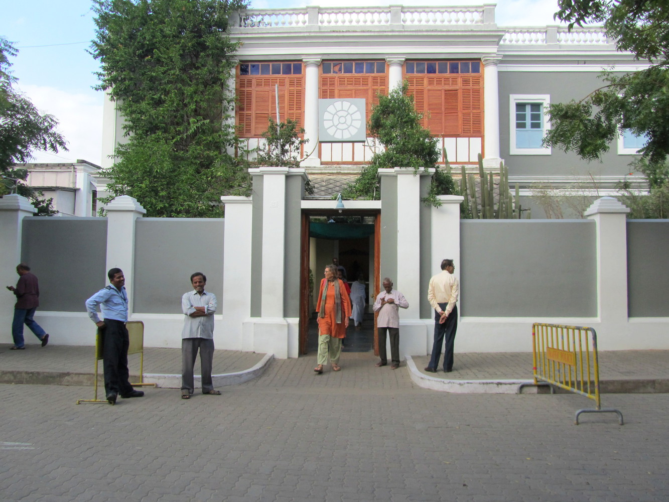 A gray, white and orange building enclosed in a gray and white compound with a gate out of which a man in orange kurta walking out