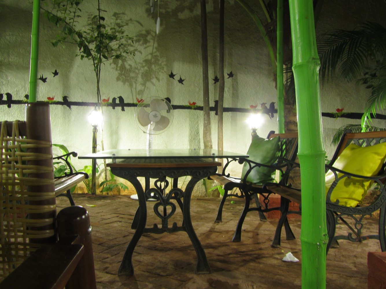 A restaurant with tables, chairs, bamboo sticks, a pedestal fan, and sketches of birds on a wall