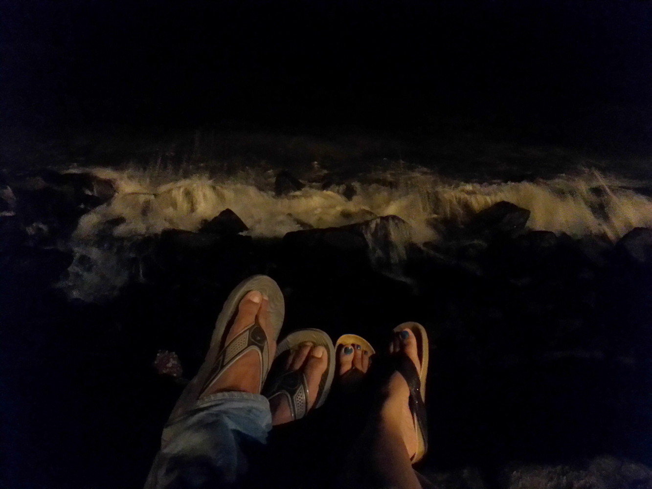 Two pairs of feet with footwear and sea waves hitting rocks in the evening