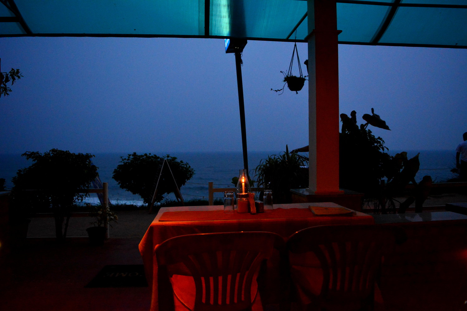 A restaurant with a candlelit table overlooking a promenade on a cliff, the sea and the dark sky at dusk